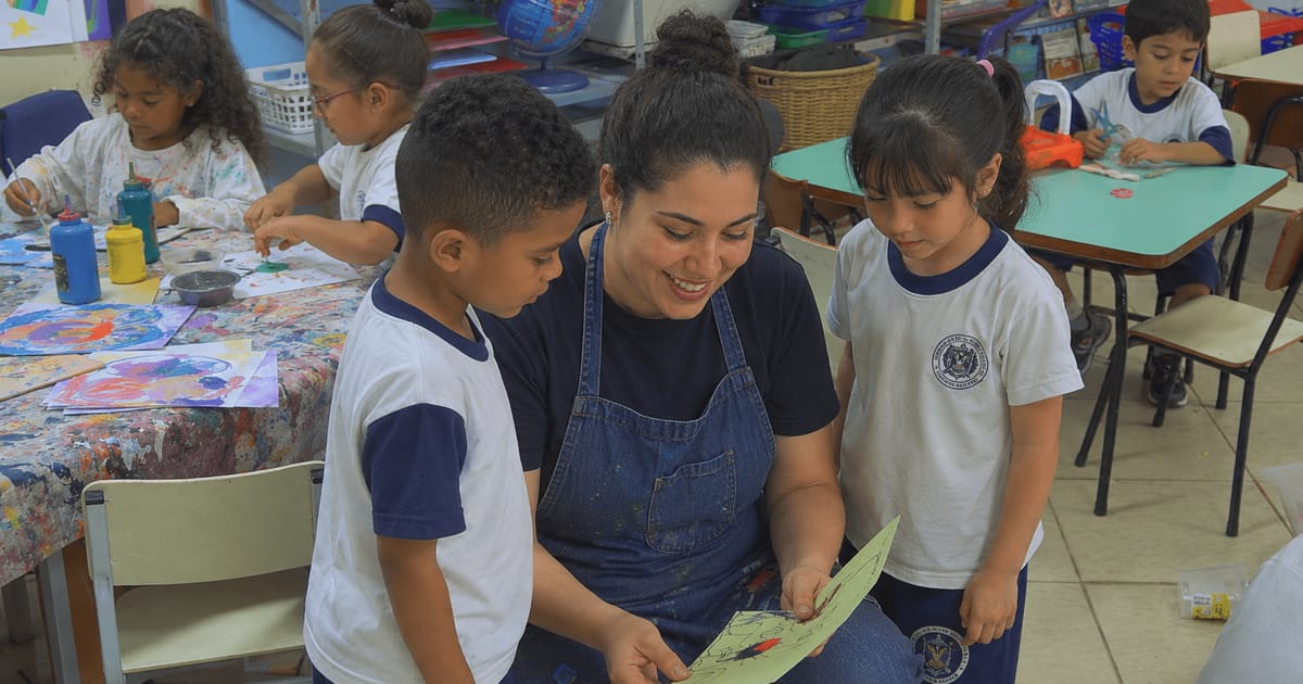 Professora sorridente orientando dois alunos em uma atividade de pintura em sala de aula.