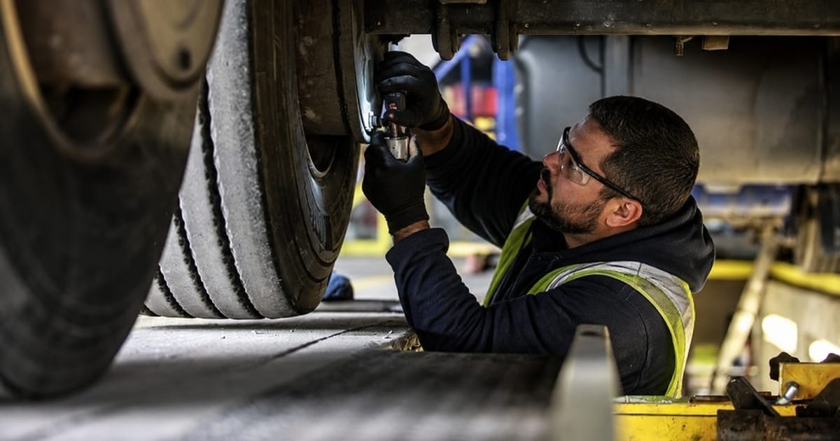 Mecânico usando óculos de proteção e luvas realizando manutenção no sistema de freios de um caminhão em uma oficina.