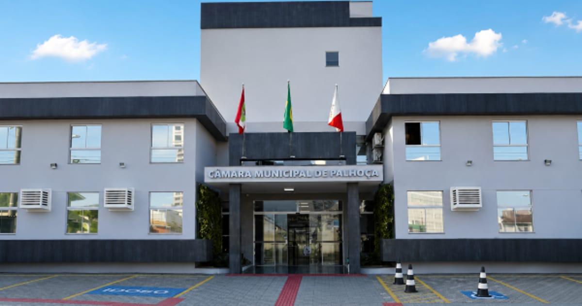 Fachada do prédio da Câmara Municipal de Palhoça com bandeiras na entrada e estacionamento com vagas reservadas.
