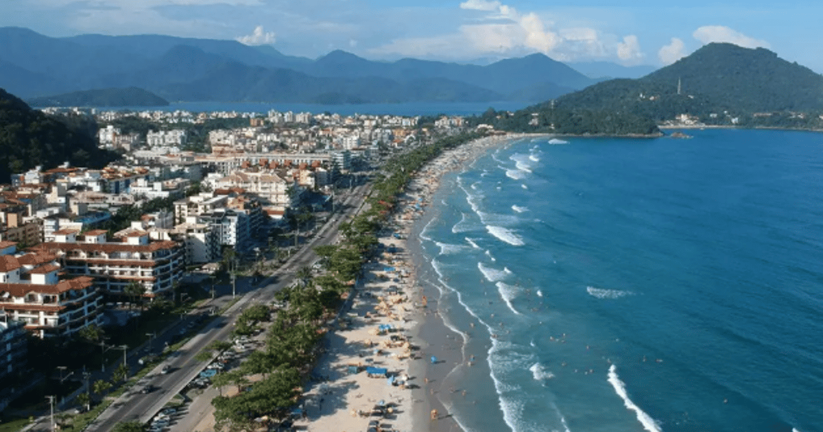 Vista aérea da orla de uma praia urbana com prédios, faixa de areia movimentada e mar azul, cercada por montanhas ao fundo.