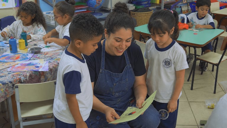 Professora sorridente orientando dois alunos em uma atividade de pintura em sala de aula.