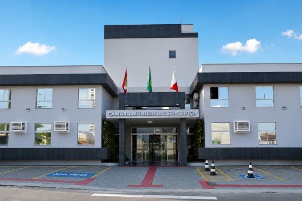 Fachada do prédio da Câmara Municipal de Palhoça com bandeiras na entrada e estacionamento com vagas reservadas.