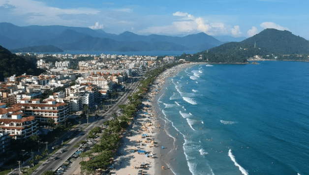 Vista aérea da orla de uma praia urbana com prédios, faixa de areia movimentada e mar azul, cercada por montanhas ao fundo.