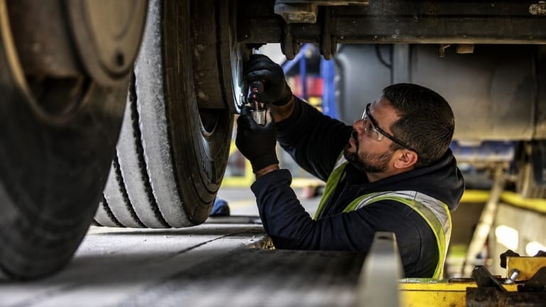 Mecânico usando óculos de proteção e luvas realizando manutenção no sistema de freios de um caminhão em uma oficina.