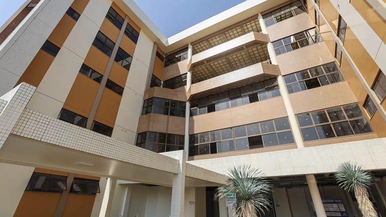 Fachada de prédio institucional com estrutura em tons de bege e marrom, janelas escuras e duas plantas ornamentais na entrada.