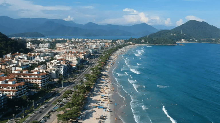 Vista aérea da orla de uma praia urbana com prédios, faixa de areia movimentada e mar azul, cercada por montanhas ao fundo.